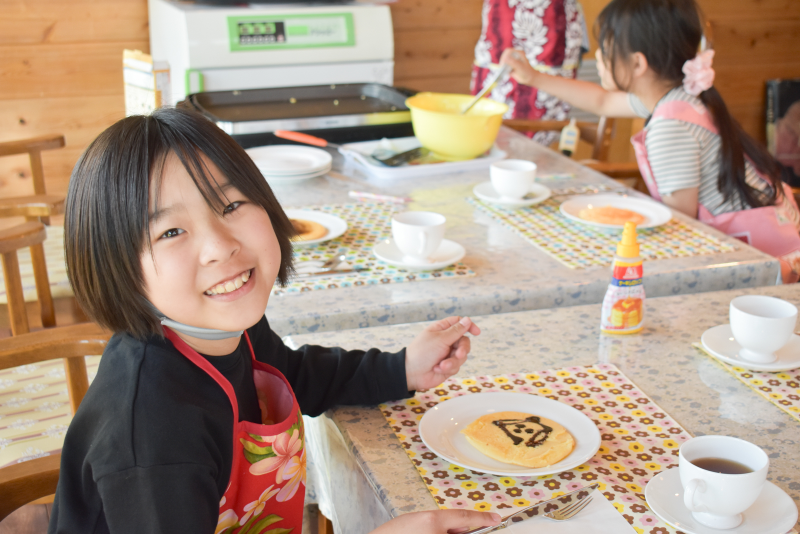 Smiling girl in a floral apron sits at a sunlit table with pancakes and tea, ready to eat breakfast.