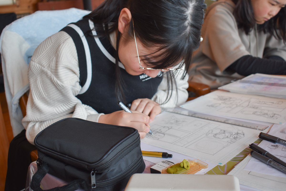 BSC_7745 | 滋賀・琵琶湖の自然体験学習施設 BSCウォータースポーツセンター Teenage student wearing glasses sketches comic panels at a desk with a pencil case and art supplies nearby.