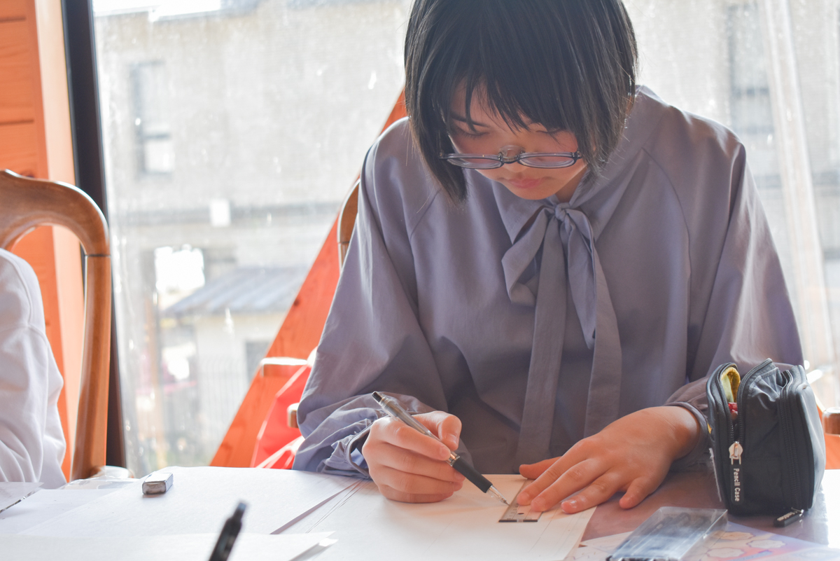 Student with glasses writes on paper at a desk using a ruler, pencil case nearby.