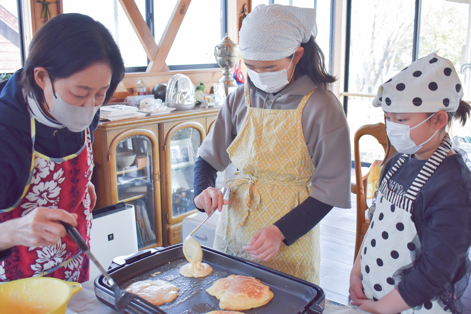 Three people in masks and aprons cook pancakes on a griddle in a cozy kitchen.