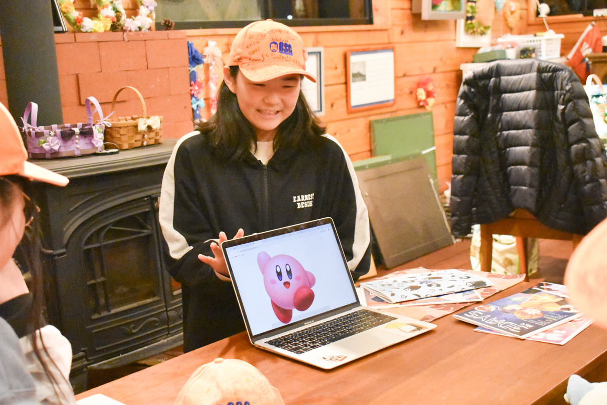 Person in an orange cap demonstrates a laptop displaying a Kirby image to a small group at a table.