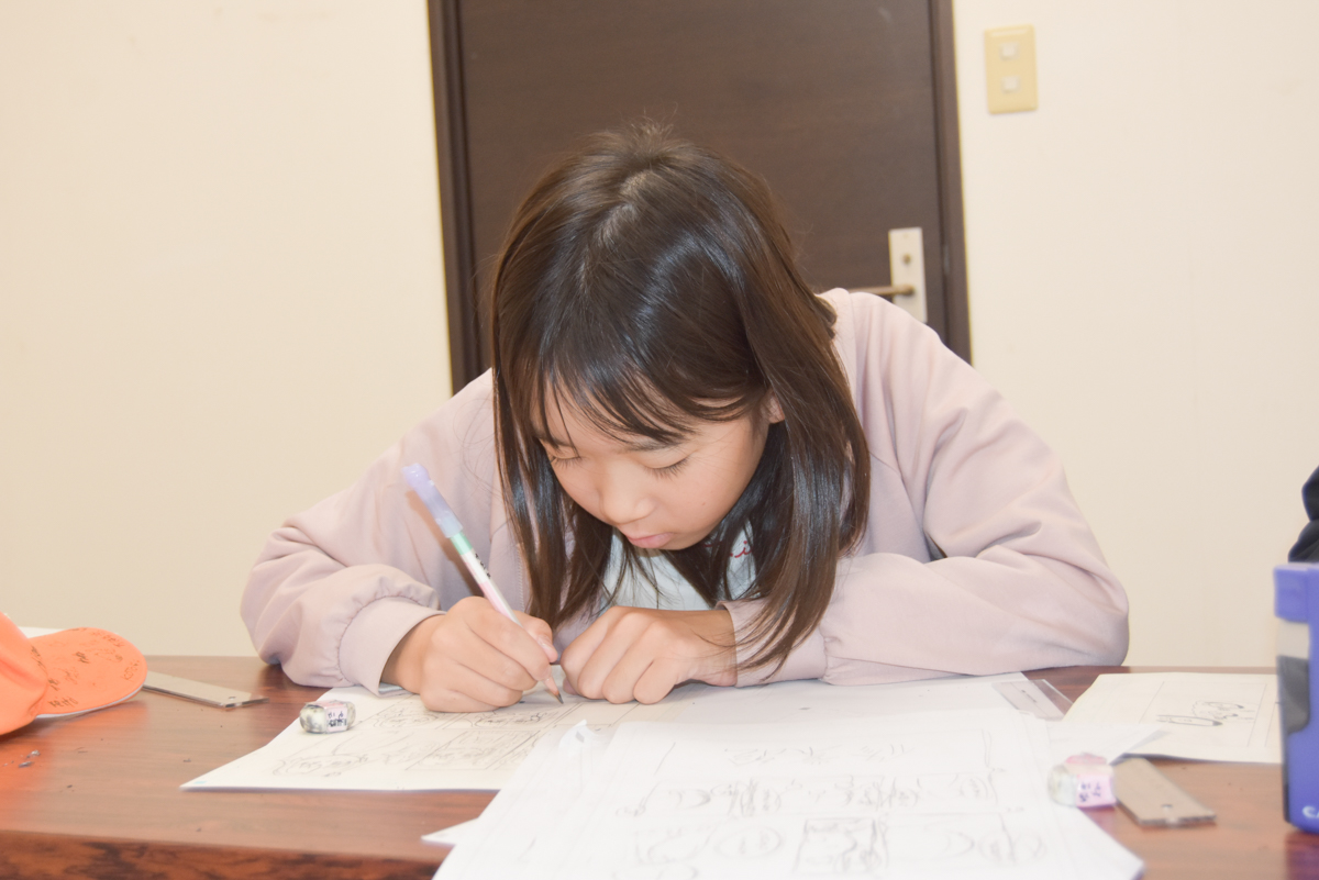 Young girl with dark hair drawing with a pencil at a wooden table, surrounded by papers and a small eraser.