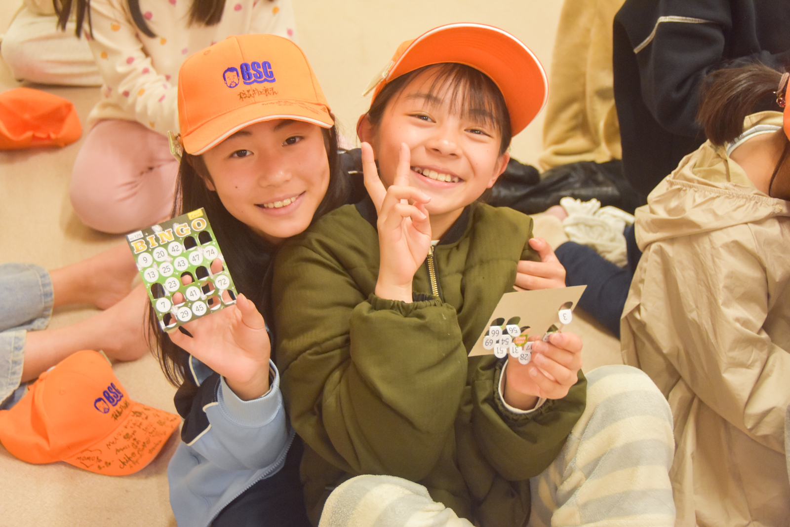 Two smiling girls wearing orange caps hold up bingo cards while sitting on the floor.