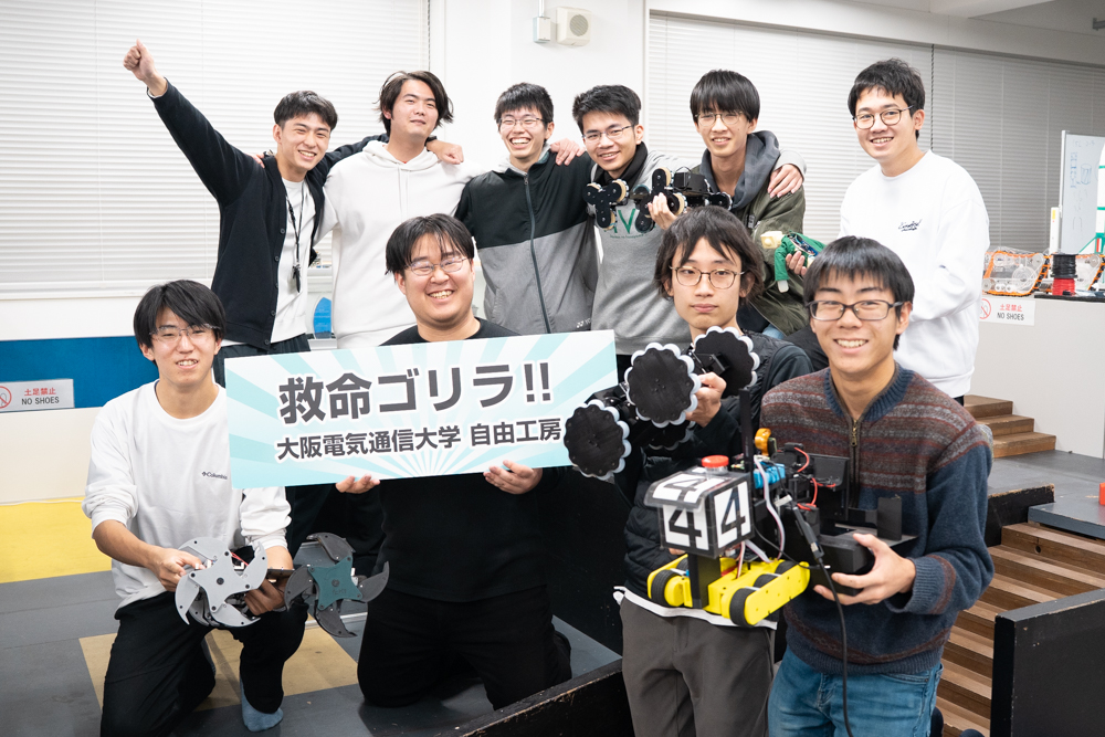Group of students in a workshop posing with a banner that reads ‘救命ゴリラ!! 大阪電気通信大学 自由工房’ and a yellow robot with the number 414 in front of them, in a lab setting.