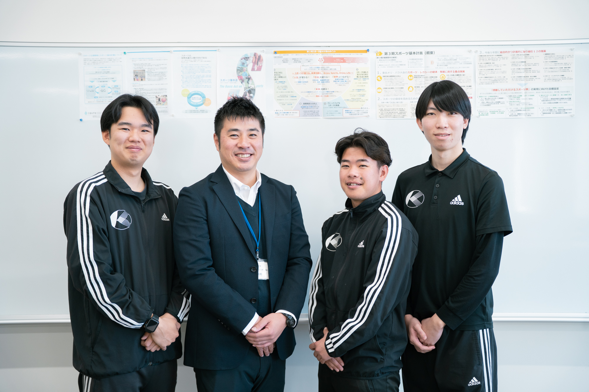 Four men pose for a group photo indoors in front of posted documents on a wall; three wear Adidas track suits, one in a suit.