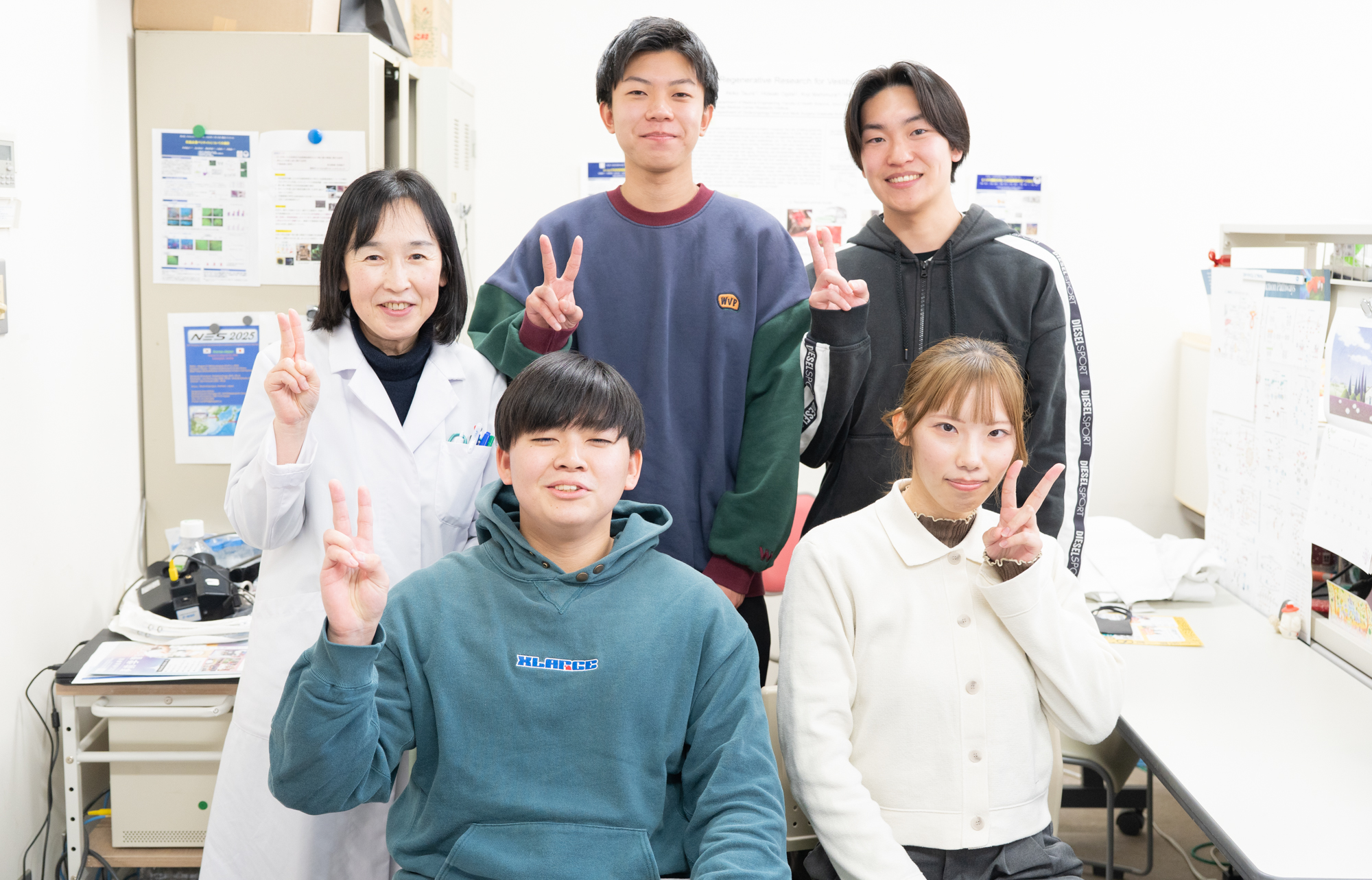 Five people—a teacher in a white coat and four students—pose in a bright science lab, all flashing peace signs at the camera.