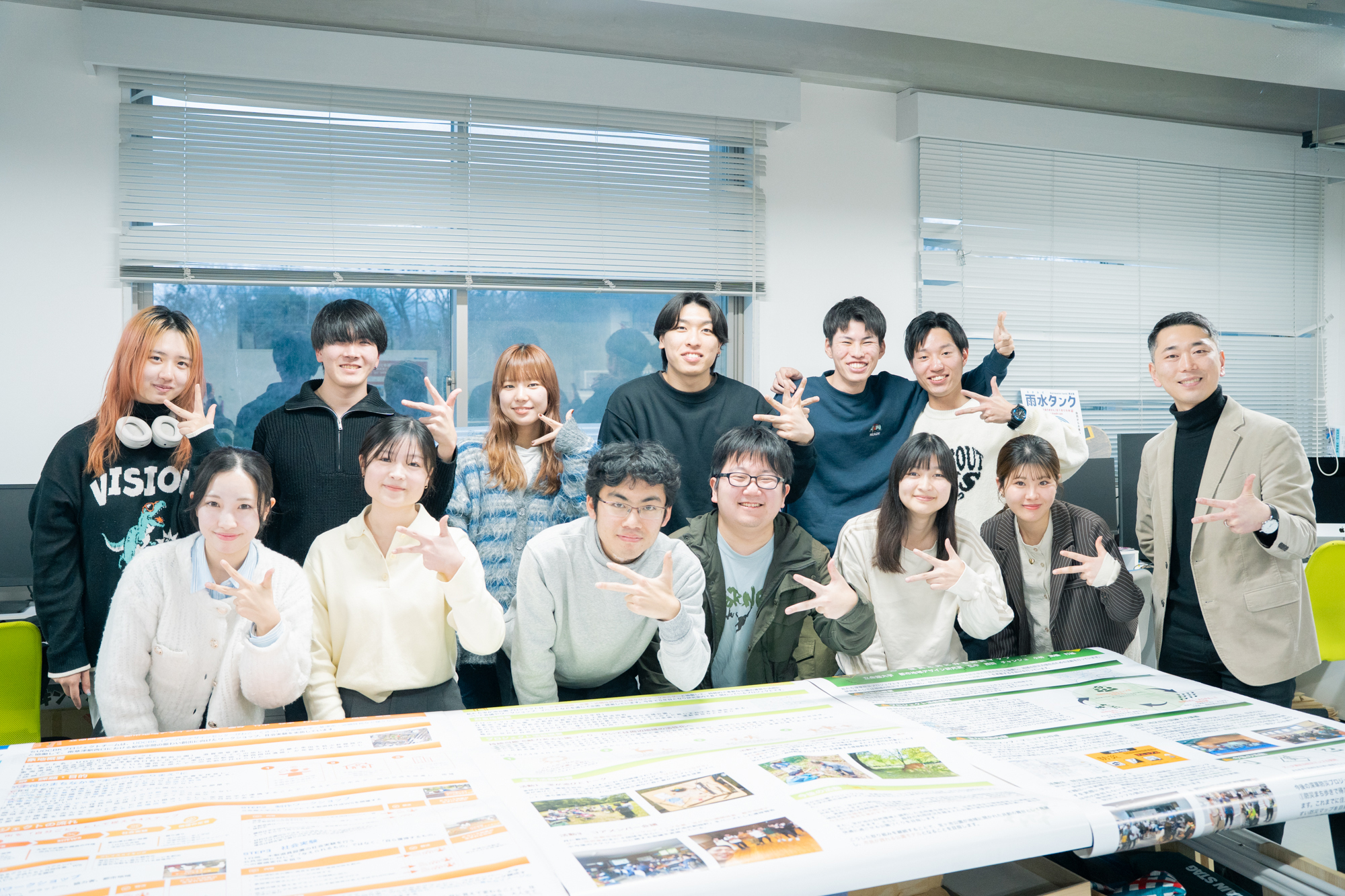 Group of students and a teacher pose for a photo in a bright classroom, standing behind a table covered with colorful brochures.