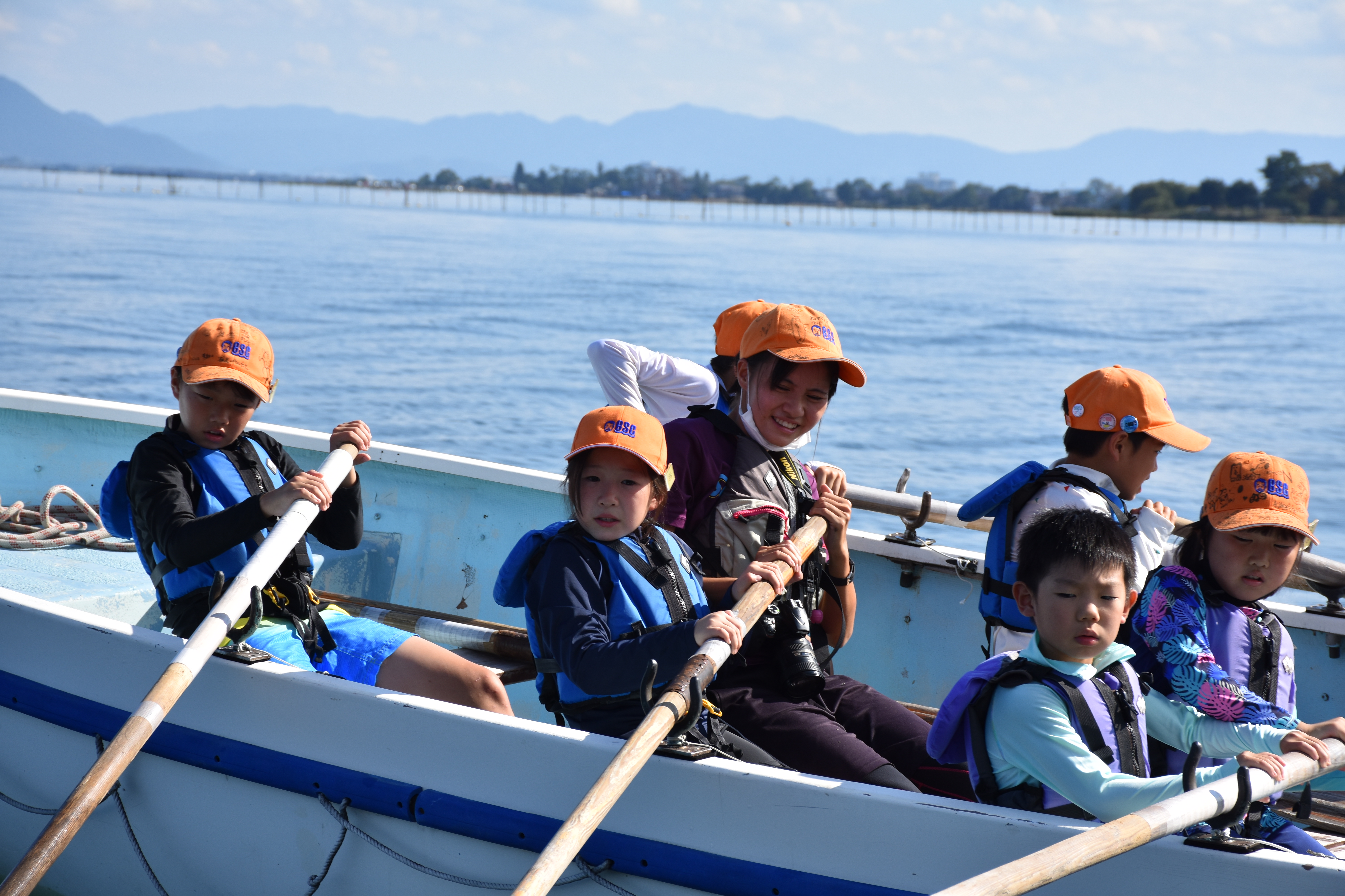 Children in orange hats and life jackets rowing a blue boat on calm water, with a distant shoreline and blue hills in the background.