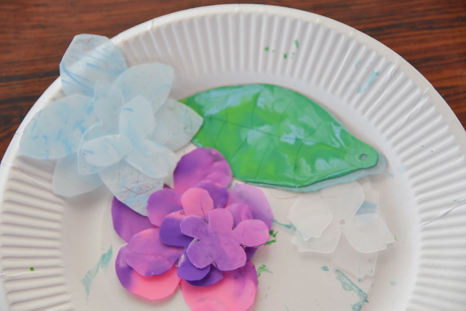 Paper plate with white and blue paper petals arranged into a floral craft beside a green plastic leaf and pink-purple flowers on the plate edge