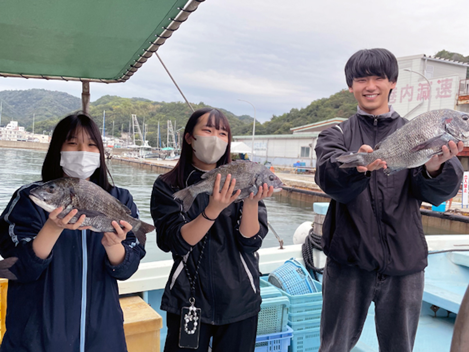 Three people wearing masks on a boat proudly hold up three large fish by the harbor with boats and hills in the background.