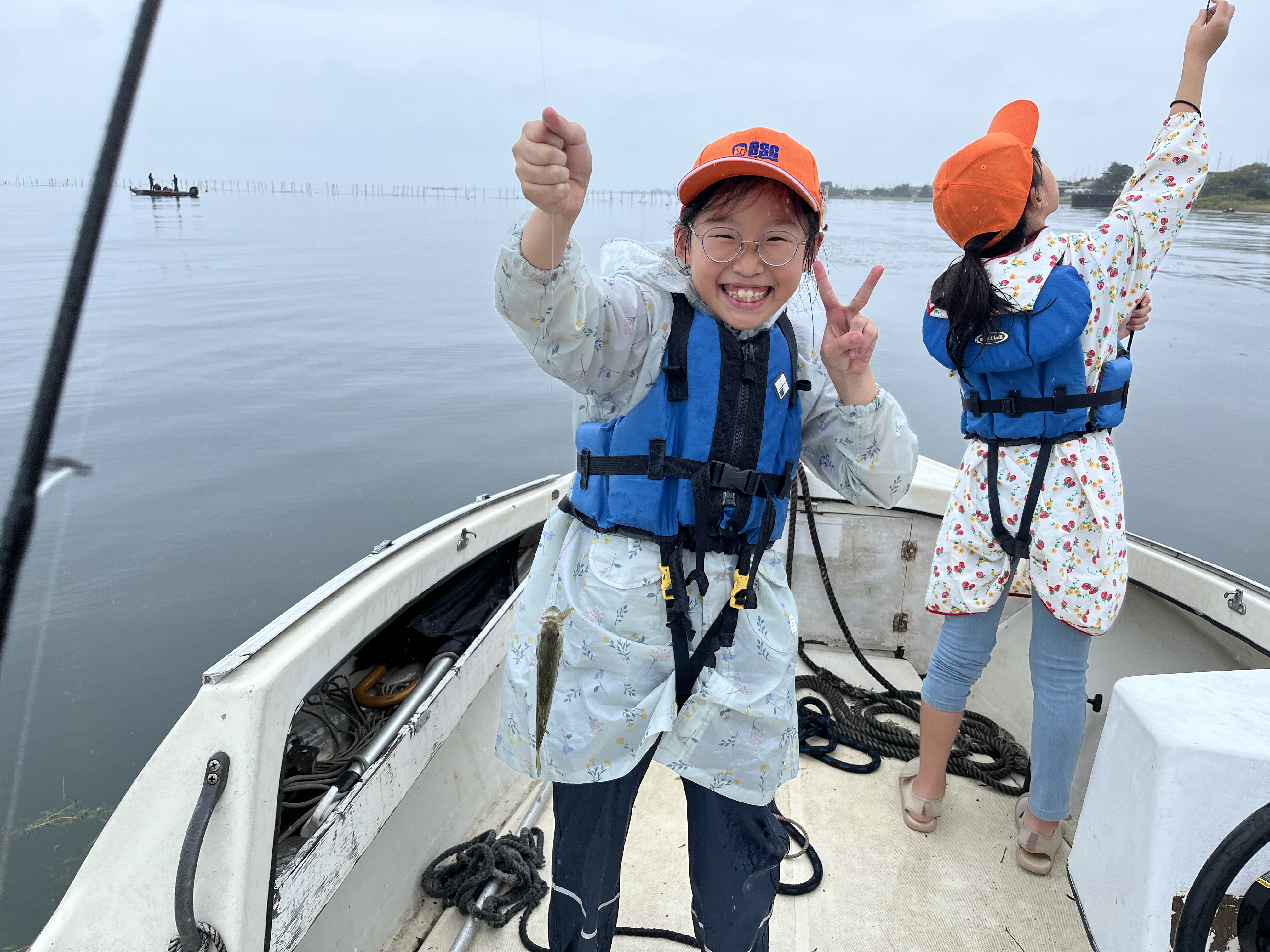 Smiling girl in a blue life jacket on a boat, holding a small fish and flashing a peace sign at the camera.