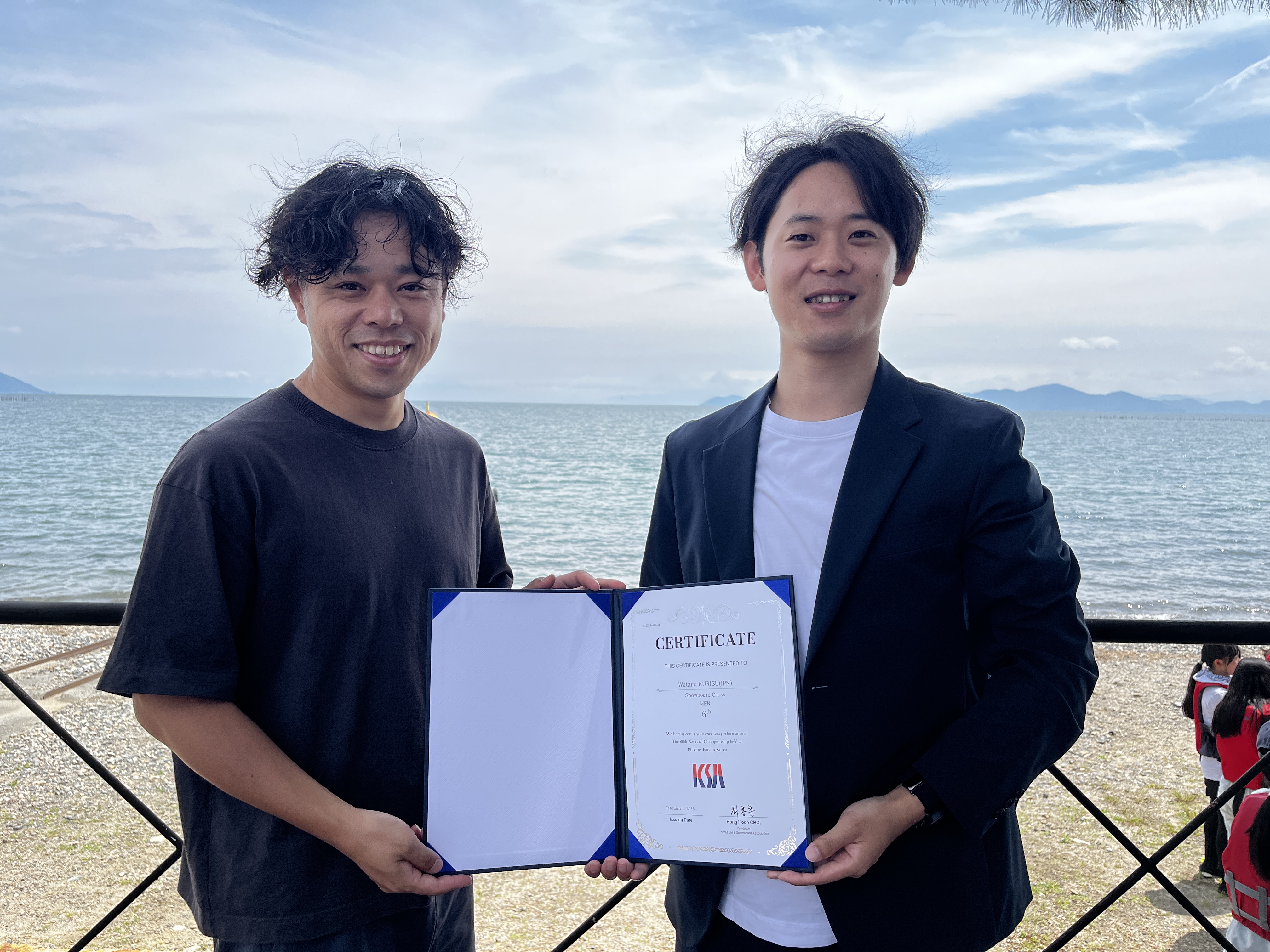 Two people stand at the seaside, smiling, while holding an open certificate folder toward the camera.