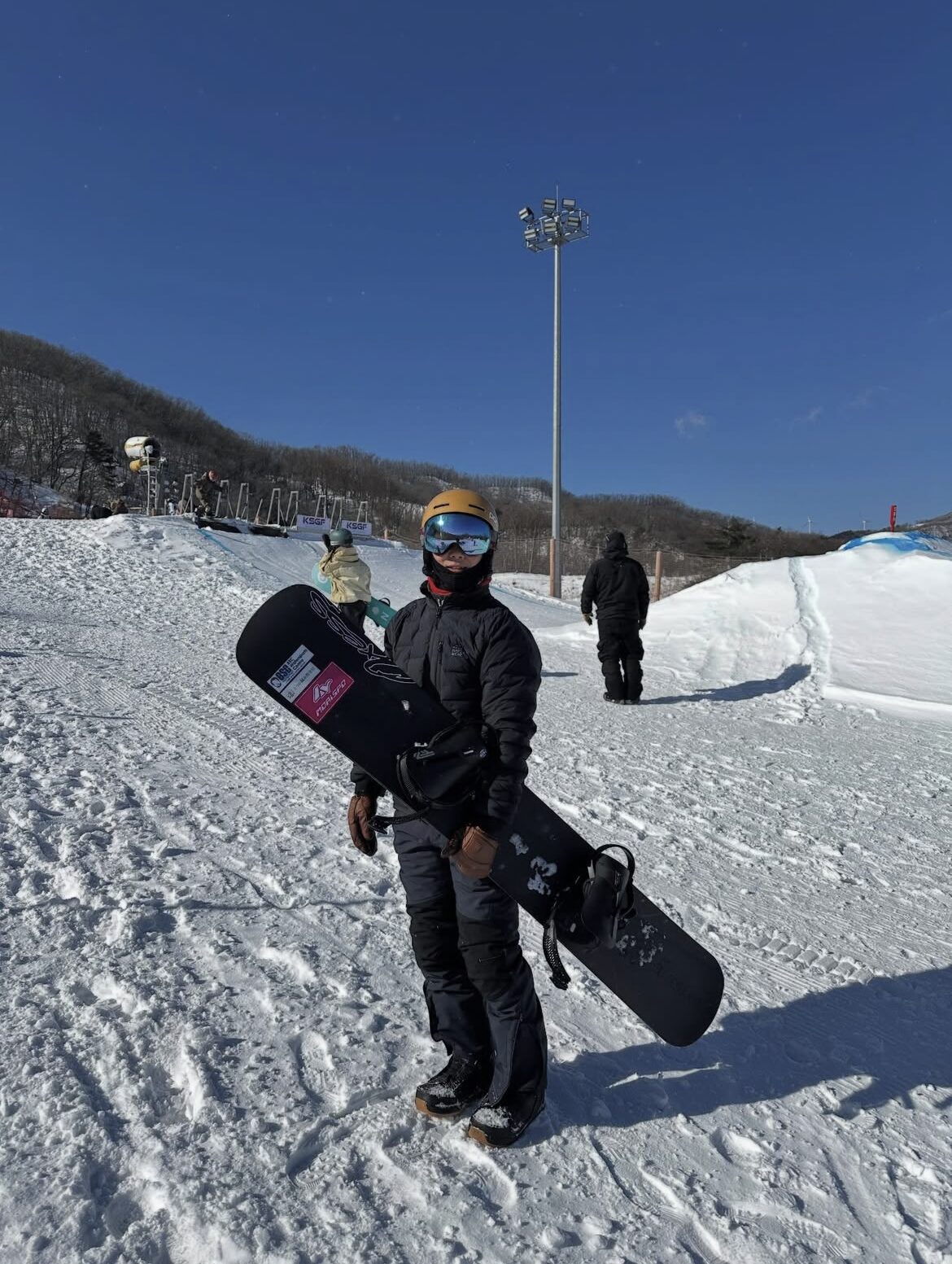 Screenshot | 滋賀・琵琶湖の自然体験学習施設 BSCウォータースポーツセンター Snowboarder in a black jacket and gold helmet holds a black snowboard on a snowy slope with a clear blue sky behind.
