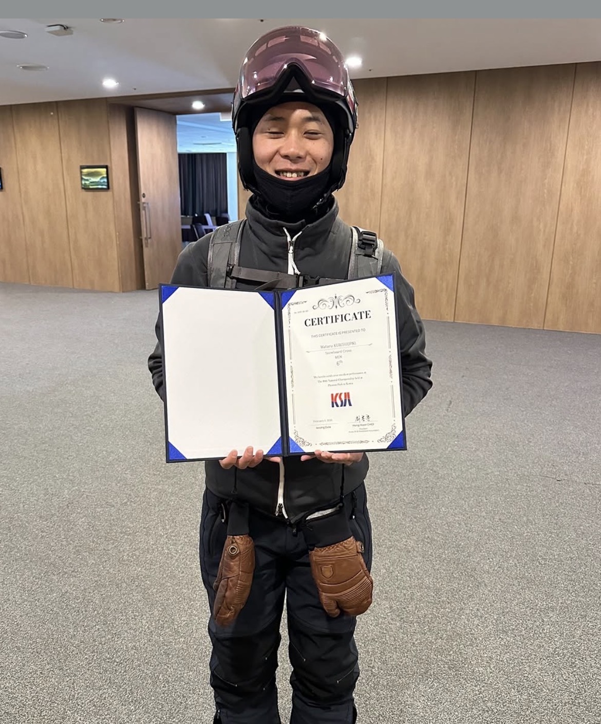Screenshot | 滋賀・琵琶湖の自然体験学習施設 BSCウォータースポーツセンター Person in ski gear and helmet holds an open certificate folder indoors, smiling at the camera.
