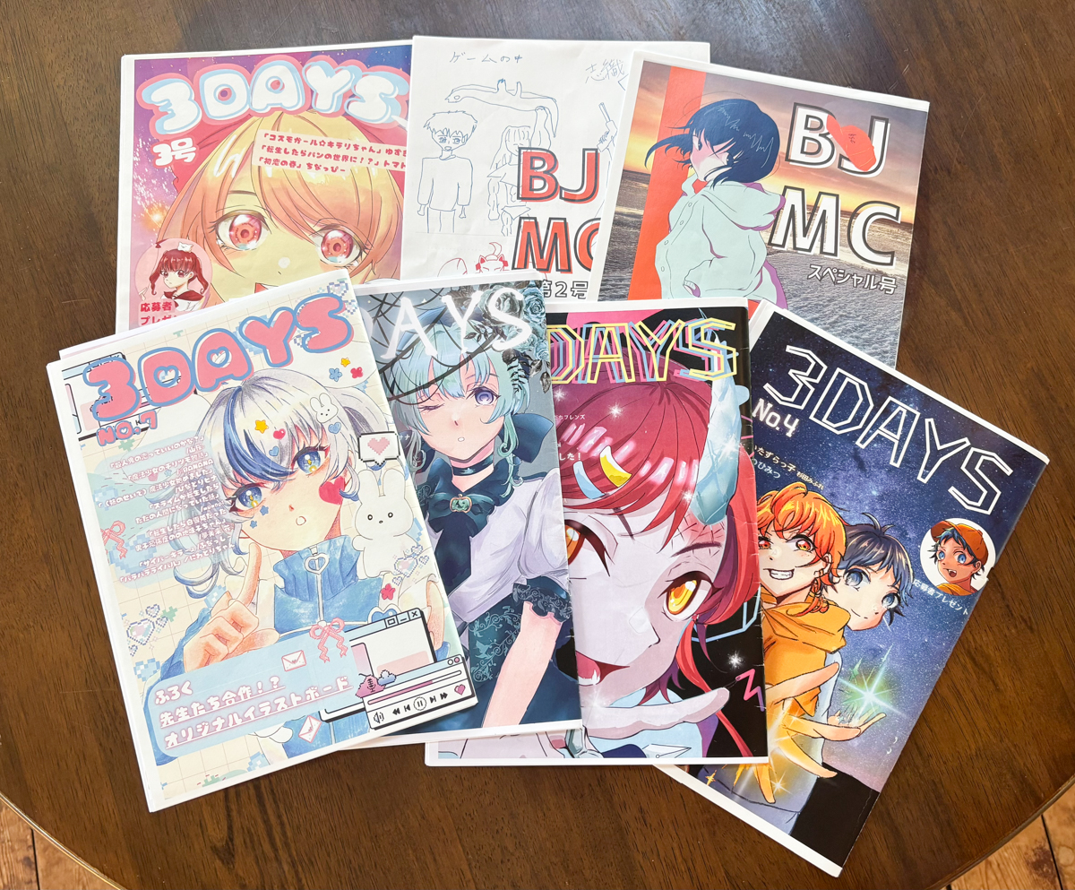 A stack of colorful Japanese manga magazines with anime-style cover art spread on a wooden table. Titles include 3DAYS and BJ MC.