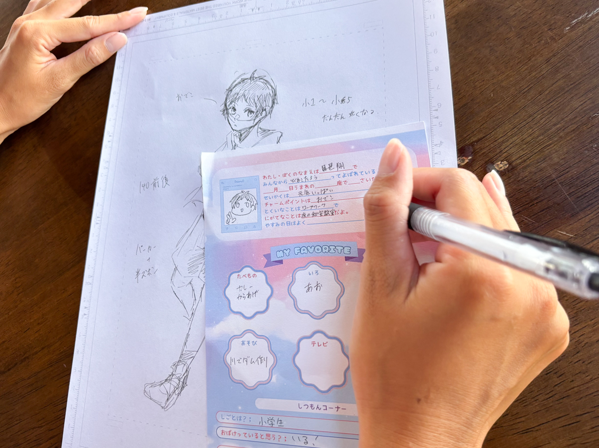 Hands sketch a manga-style character on tracing paper with a ruler along the edge, a colorful worksheet resting on a wooden table in front.