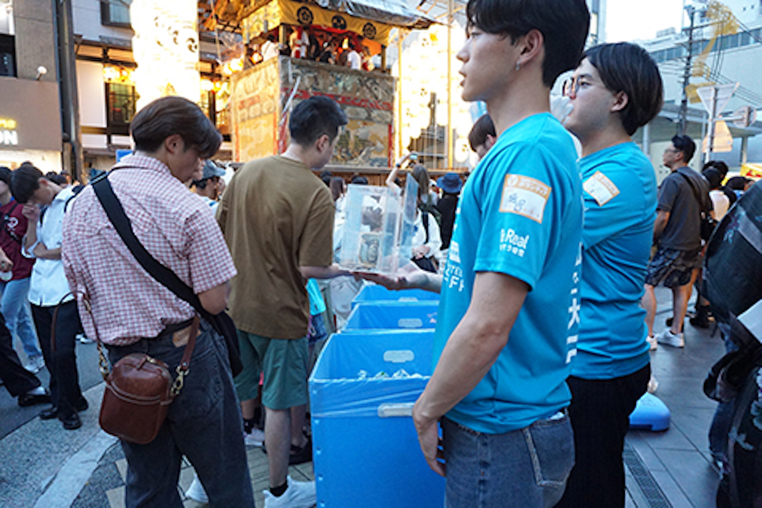 Two volunteers in blue shirts hand a clear plastic box to a crowd at a bustling street festival with a lantern float in the background