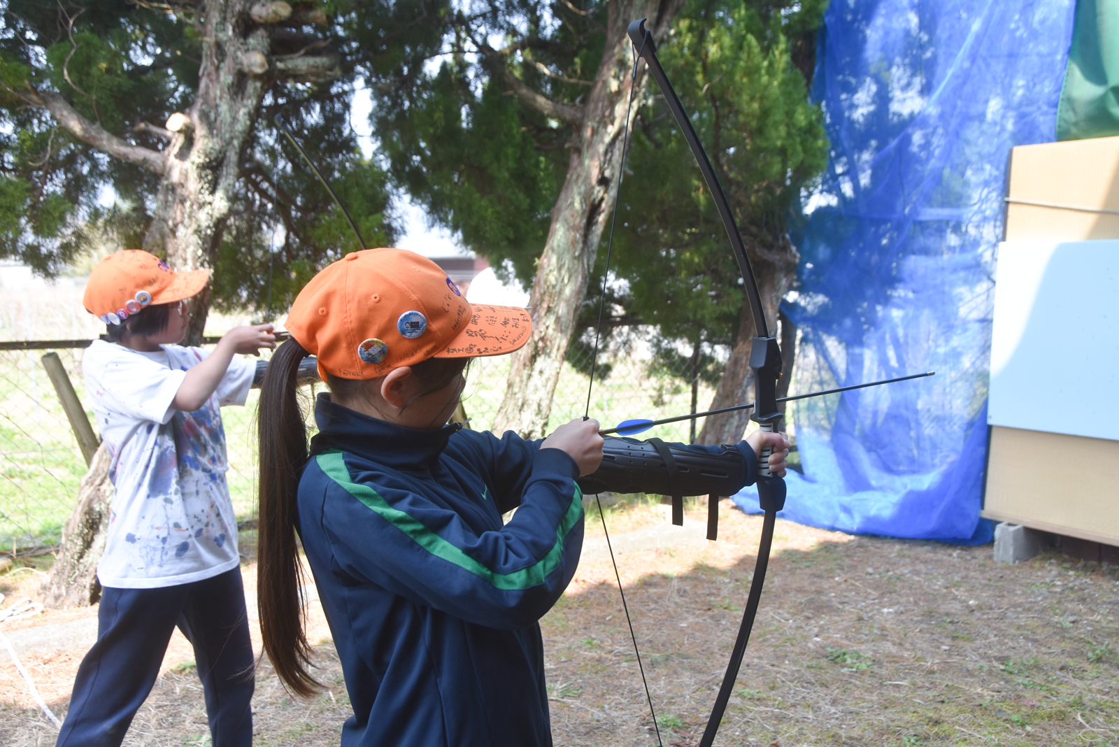 Two children practice archery outdoors, aiming with bows beside a blue netted backdrop.
