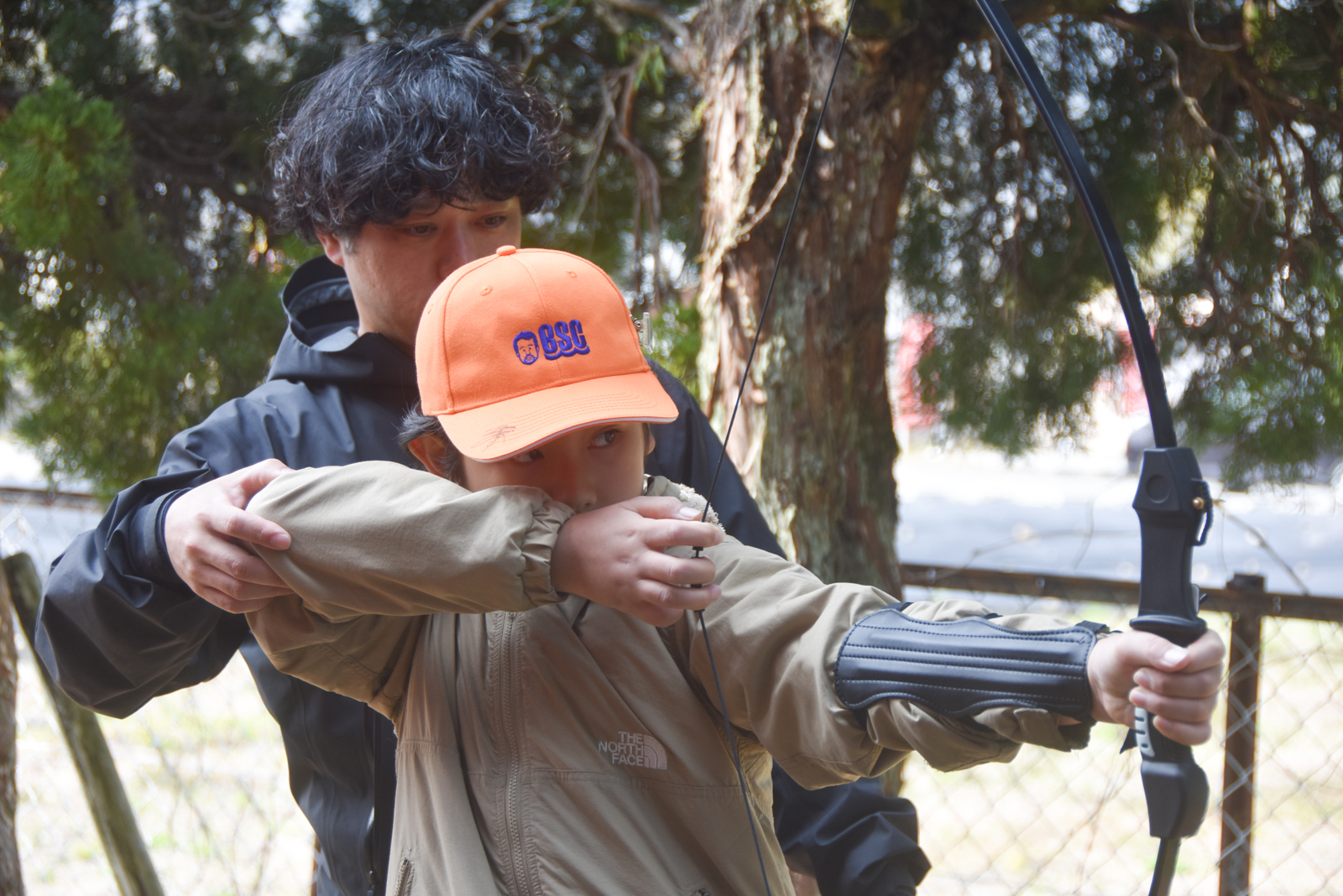 Child in an orange cap draws a bow while an instructor guides from behind during an outdoor archery lesson.