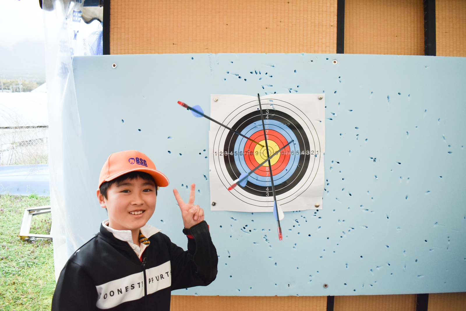 Smiling boy in an orange cap stands beside a bullseye archery target, with arrows near the center and many holes in the board behind him.