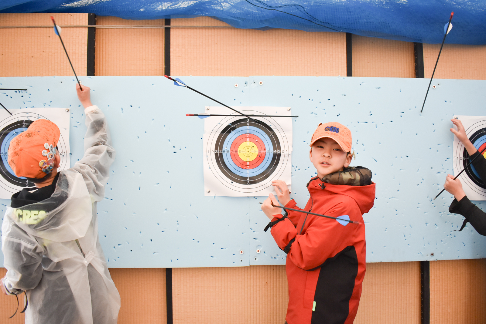 Two children practice archery at a classroom-style range, aiming at stacked targets on a blue backboard, arrows lodged in targets and on the wall nearby