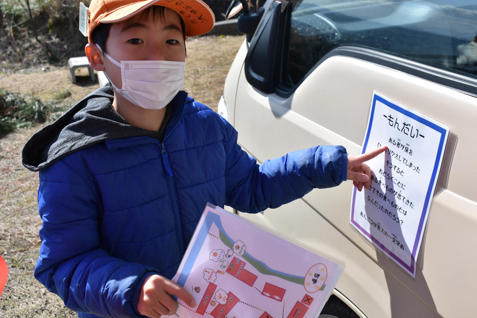 Child in an orange cap and blue jacket points to a notice posted on a beige van while holding a colorful booklet and wearing a mask.