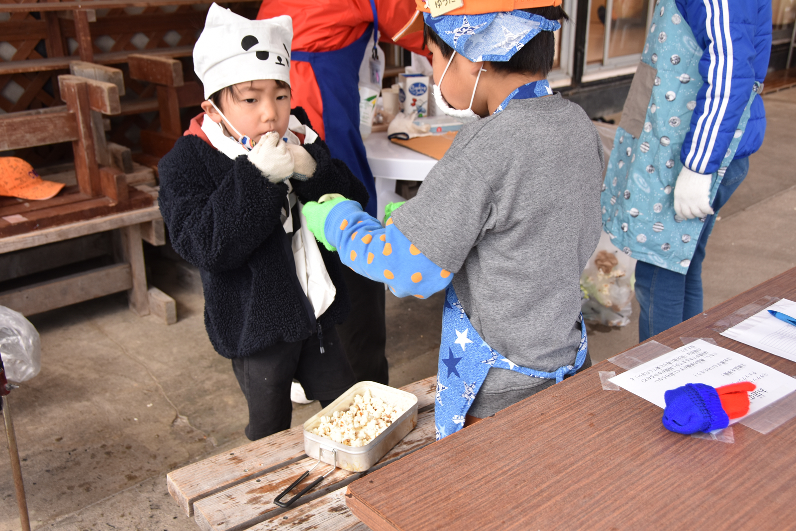 Two kids in costume at a wooden table; one hands popcorn to the other from a metal tin.