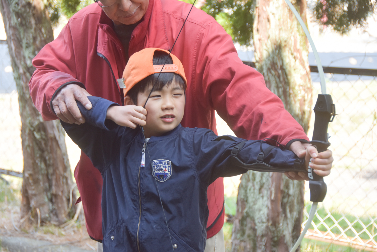 Young boy in an orange cap practices archery with a supervising adult outdoors, near a fence.