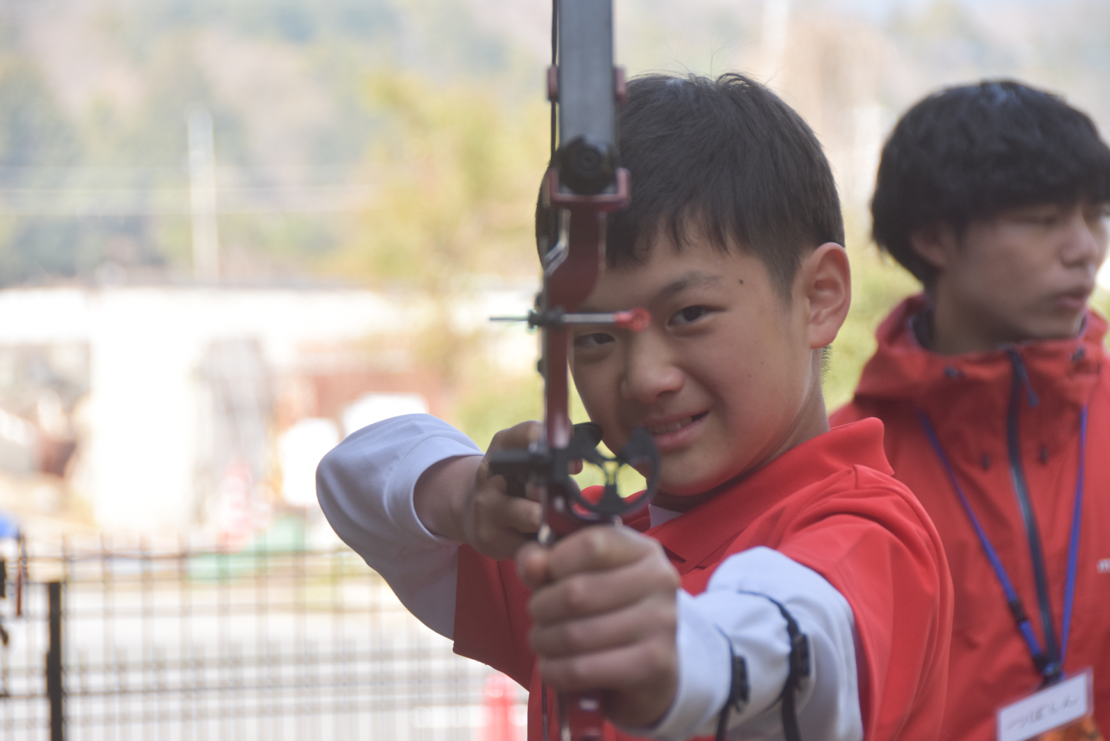 Boy in a red jacket aims a bow outdoors, focusing on the target while another person watches in the background