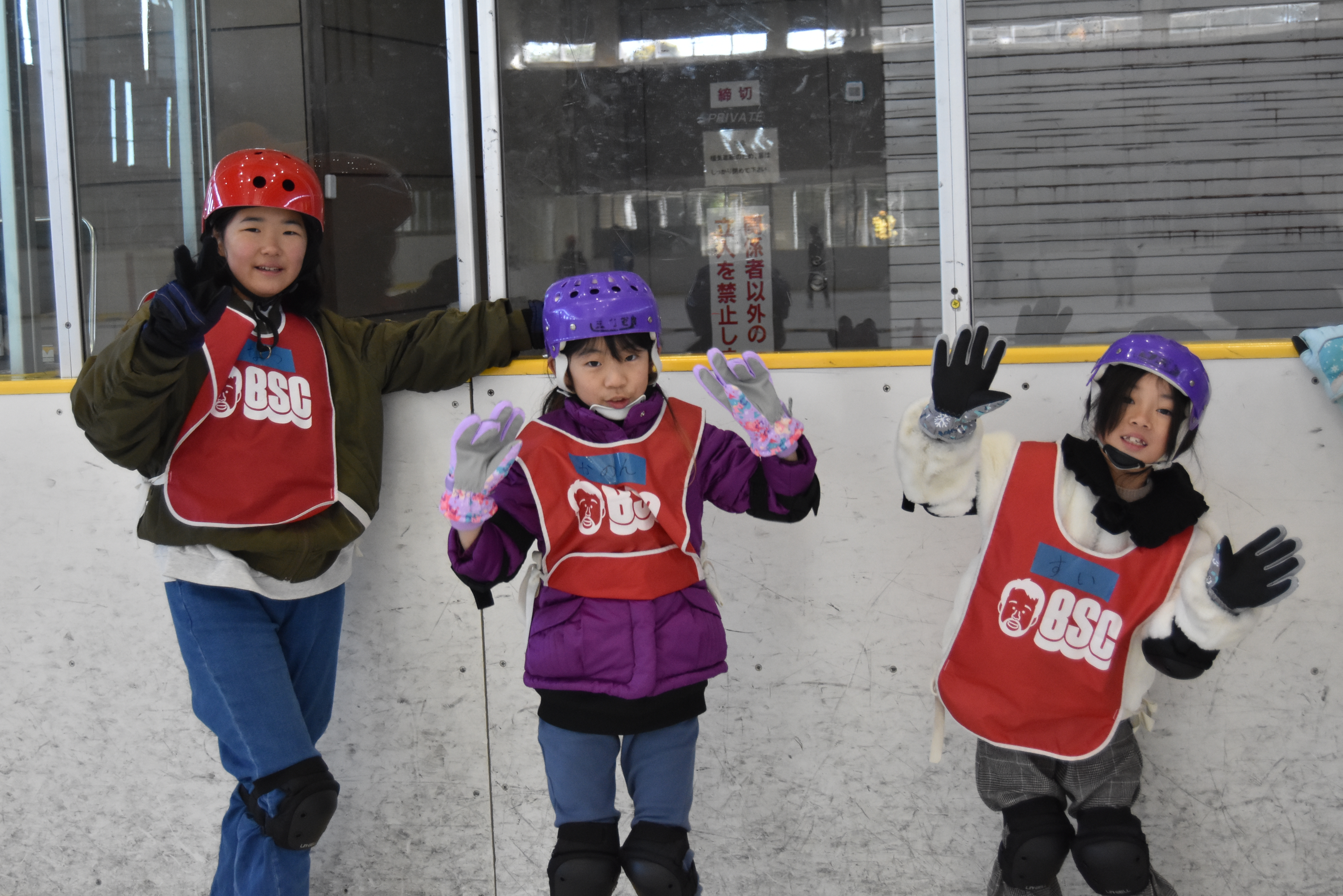 Three kids in helmets and red bibs pose and wave on an indoor ice rink, smiling at the camera.