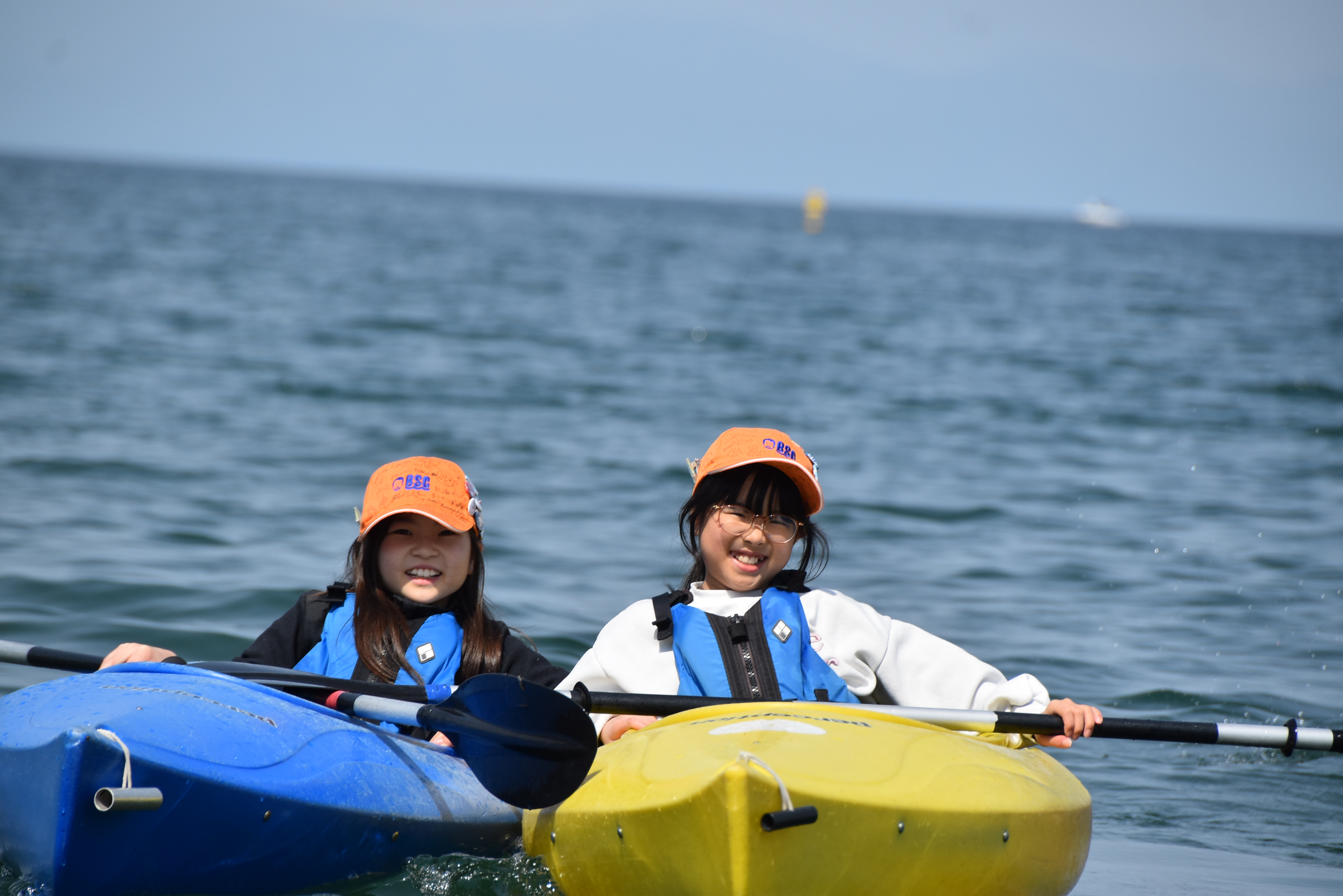 Three people wearing orange caps and blue life jackets sit in colorful kayaks on calm sea, smiling at the camera with a distant horizon behind them.
