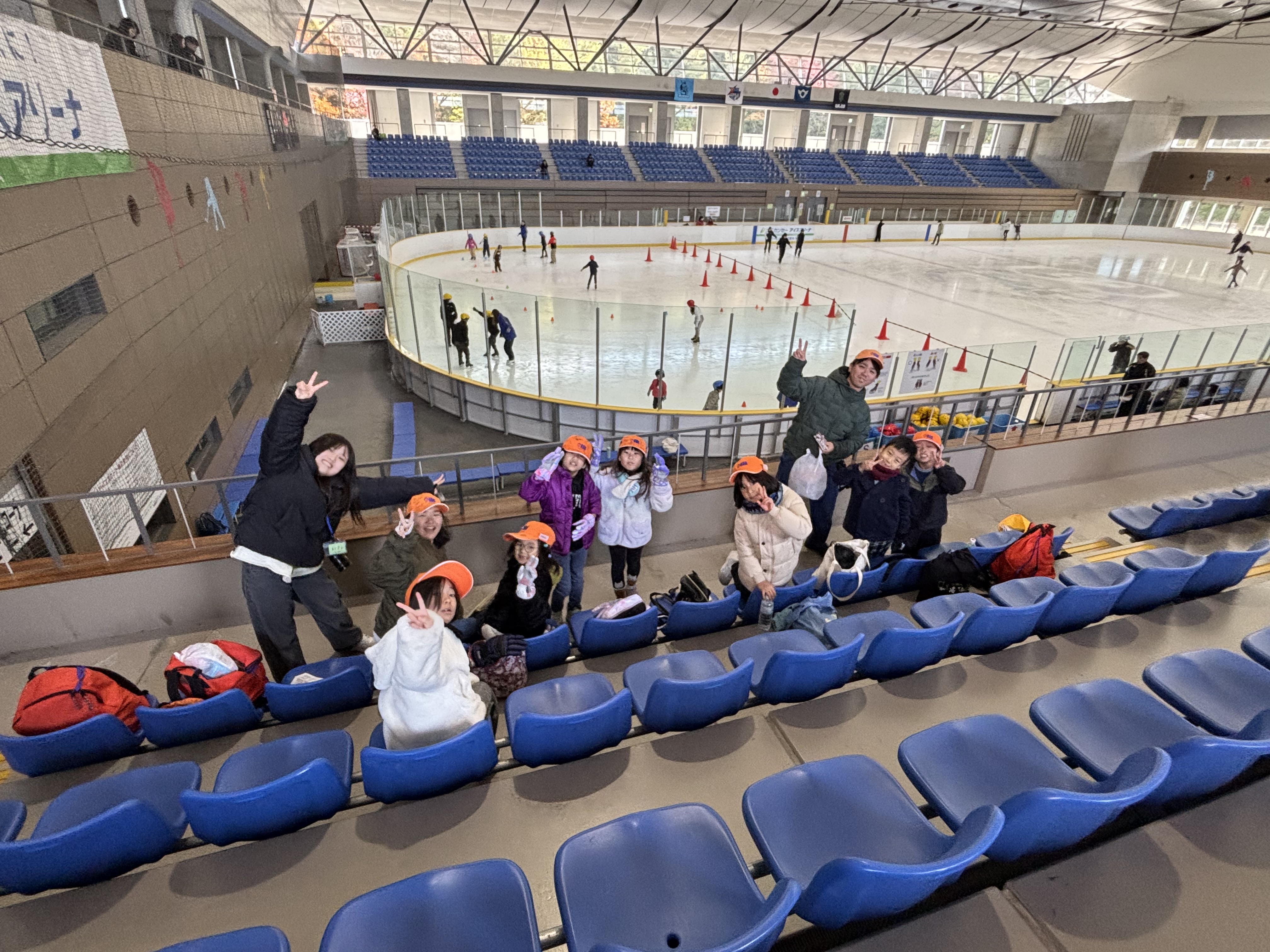 Children wearing orange hats wave from the stands as skaters practice on an indoor ice rink behind them, with blue seating above and cones marking a course beneath the glass barrier.