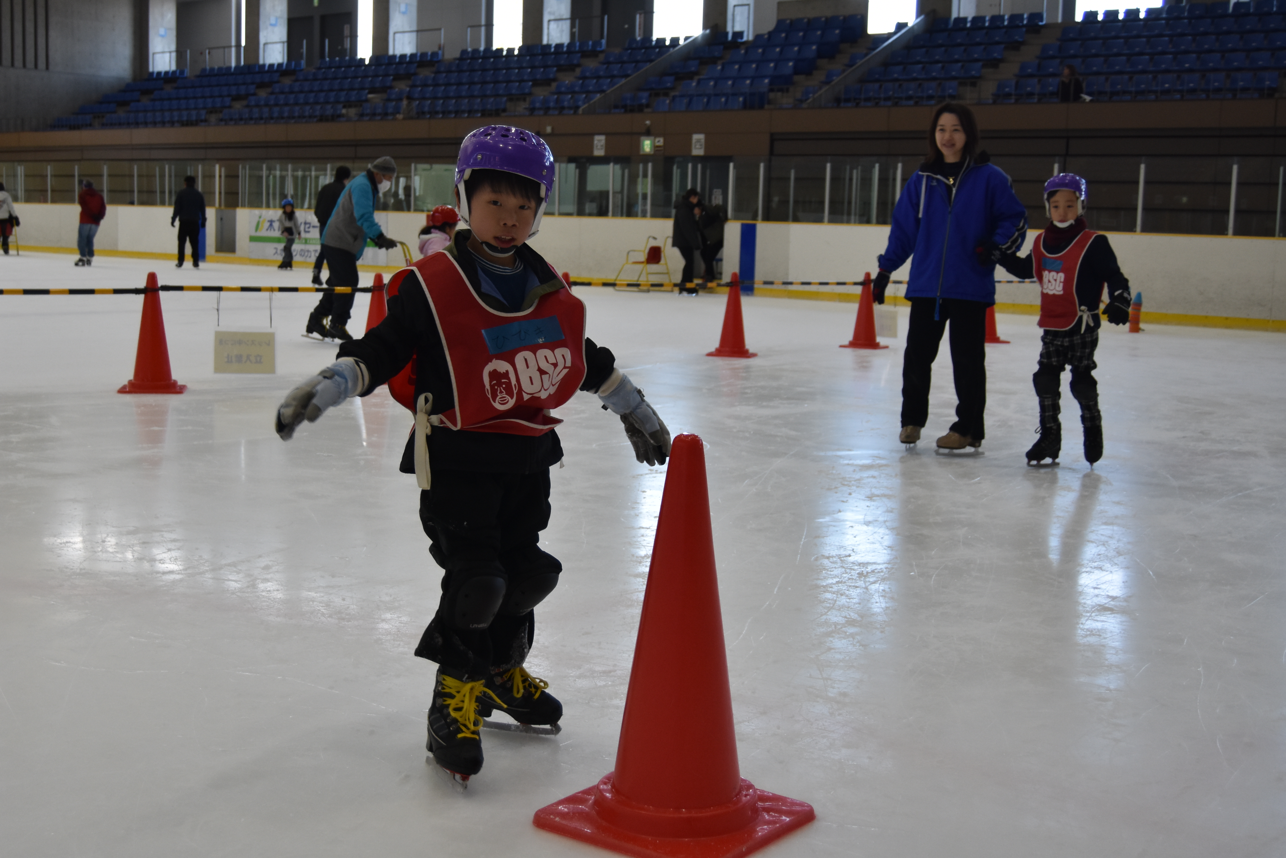 Child in a purple helmet and red vest skates around an orange cone on an indoor ice rink, with a supervising adult and other skaters in the background.
