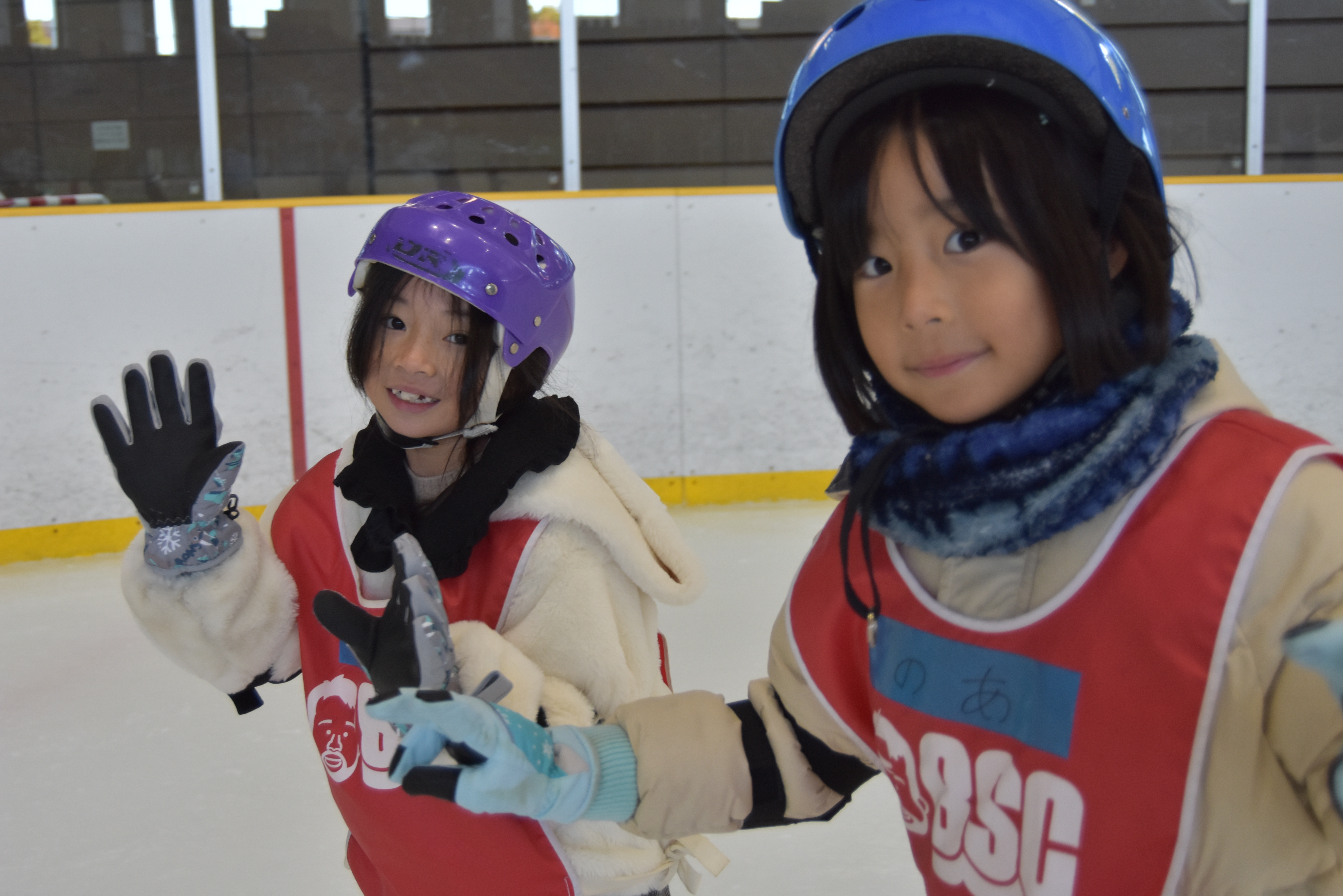 Two young girls in helmets and red vests on an ice rink, waving at the camera.