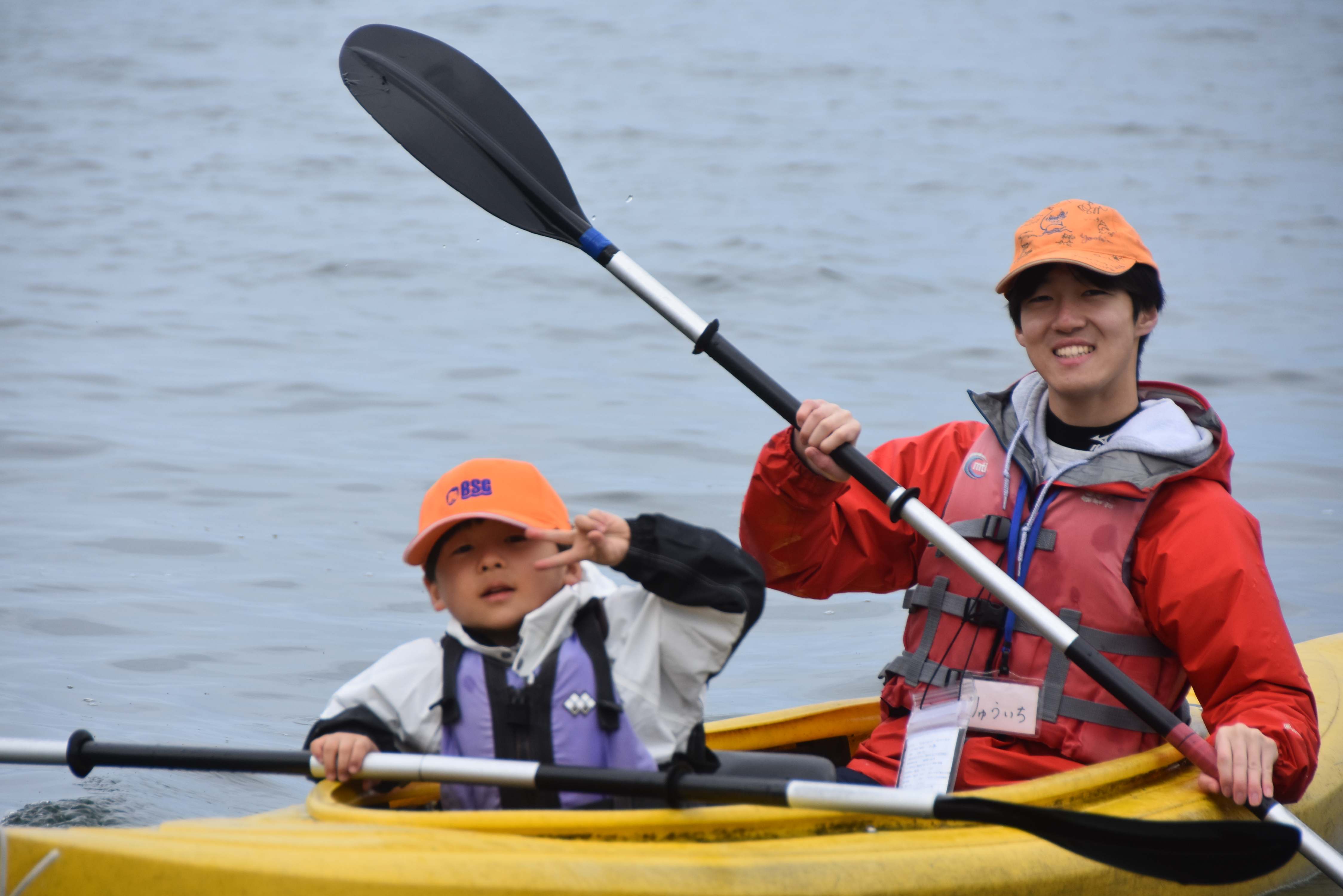 Two people in a yellow kayak paddling on calm water, wearing life jackets and bright hats (adult on right, child on left).