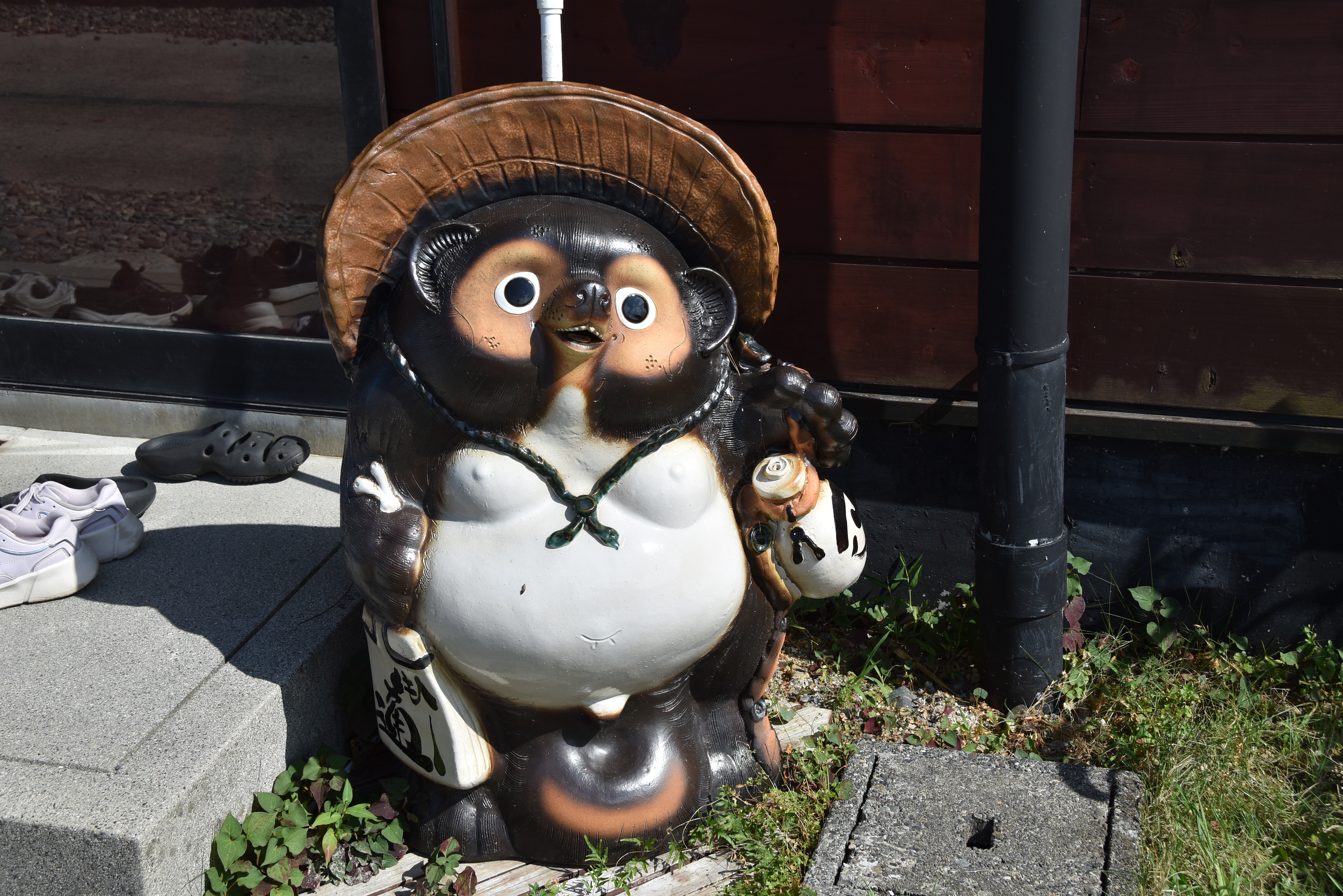 Decorative tanuki statue with a large hat on a sidewalk near a wooden wall and a black drainpipe.