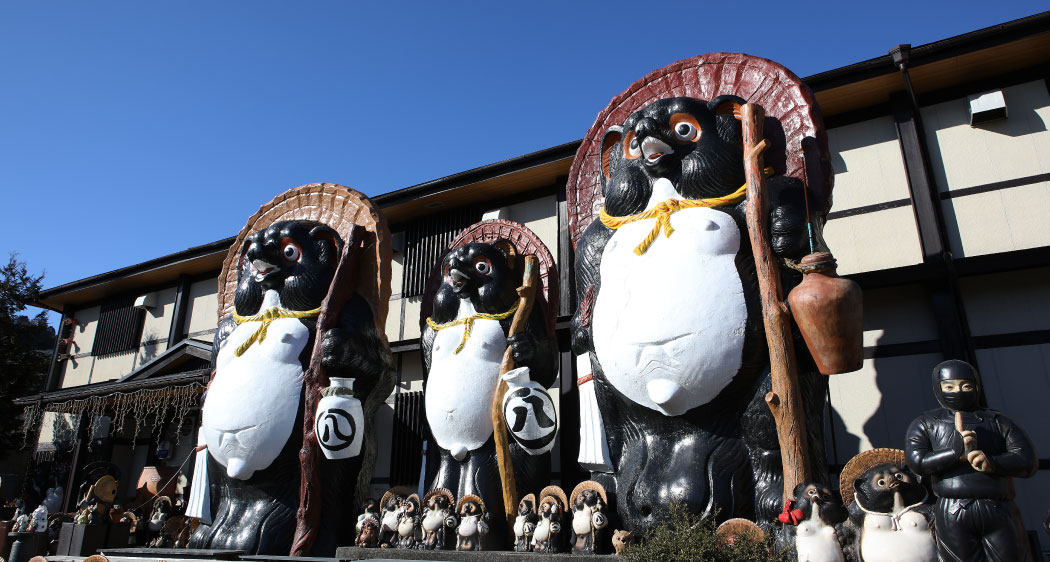 Three large tanuki statues with gourds and staffs outside a shop, surrounded by many smaller tanuki figures under a blue sky.