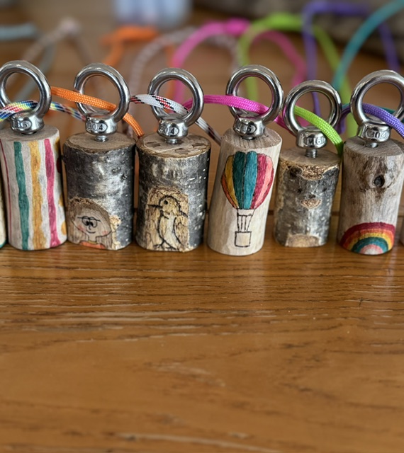 Row of wooden charms with metal rings and colorful cords, each decorated with different hand-drawn designs (rainbow, balloon, figures).